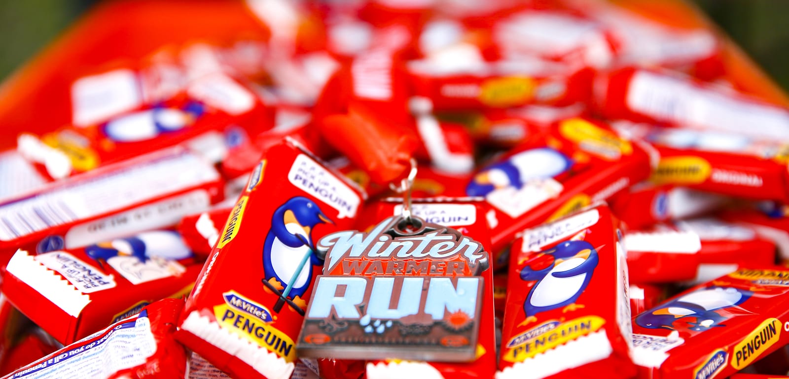 A close-up of numerous red and white candies featuring a penguin character, surrounding a "Winter Warmer Run" medal. The colorful wrappers and metallic badge create a vibrant and festive scene.