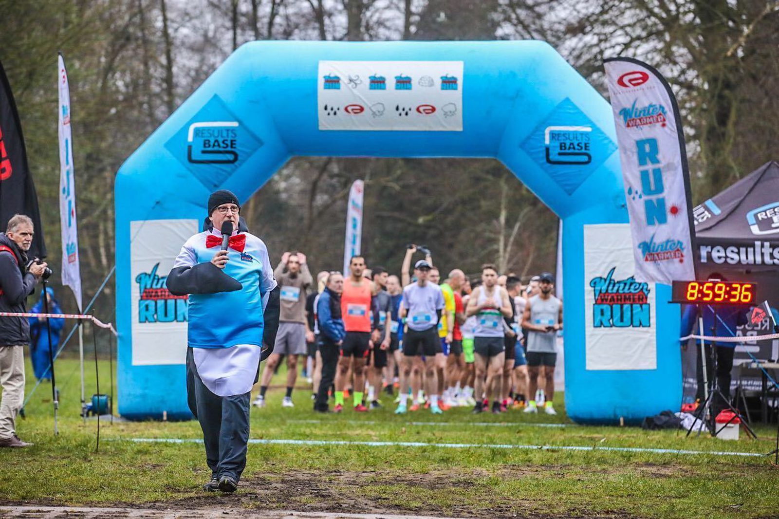 A person in a costume stands at a race starting line, holding a microphone. Behind them, a group of runners is preparing to start. An inflatable archway marked with "Winter Wonderland Run" and other sponsor logos frames the starting area. A clock displays 8:59:36.