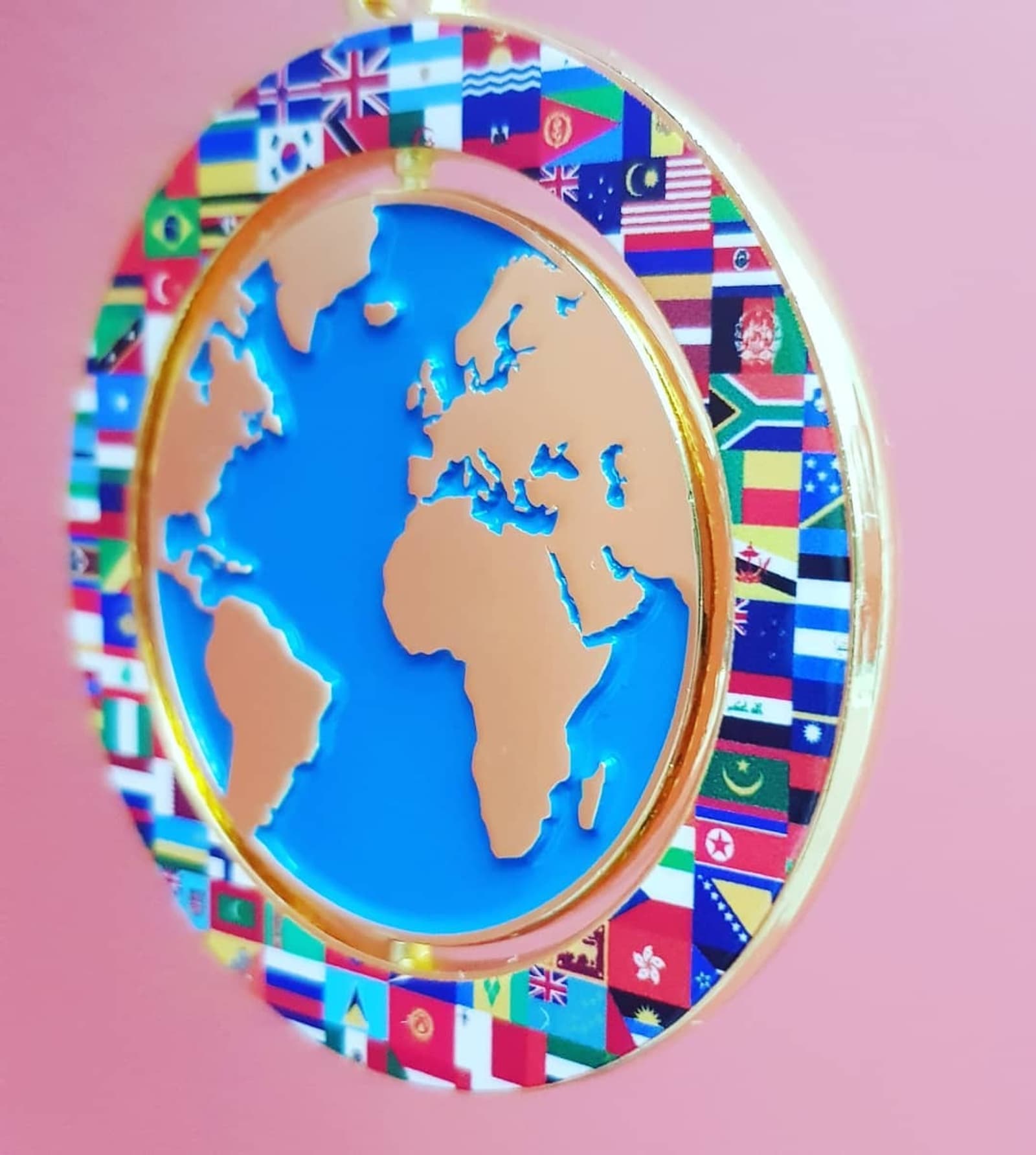 A 3D depiction of a world map in blue and copper tones is encircled by various international flags against a soft pink background. The circular frame consists of multiple small flags representing different countries from around the globe.