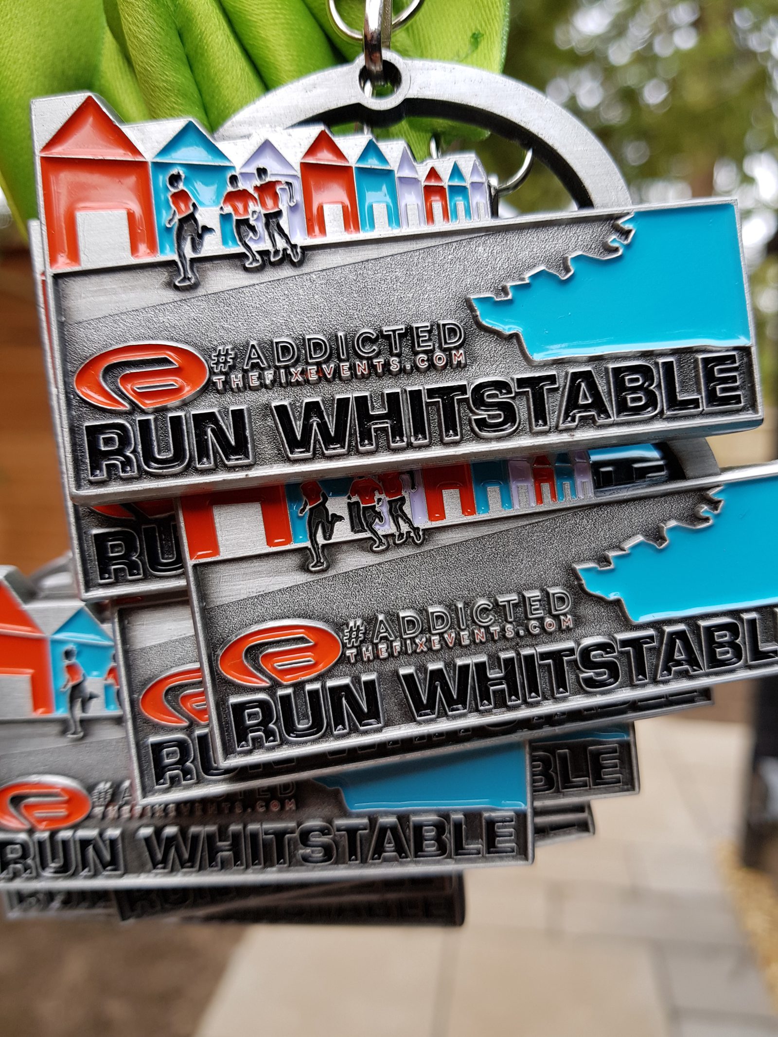 A close-up shot of multiple metallic medals hanging on green ribbons. The medals feature colorful houses, small runner figures, and the text “RUN WHITSTABLE” prominently. The background is blurred, hinting at an outdoor setting with a pathway and trees.