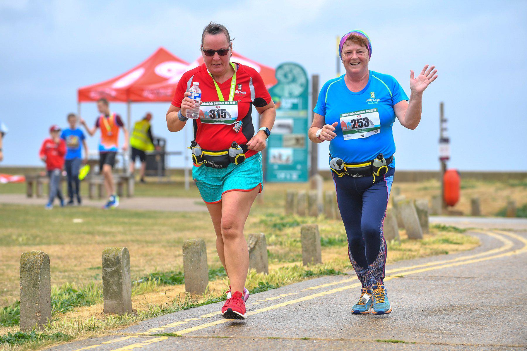 Two women participating in a race run on a paved path outdoors. The woman on the left wears a red shirt, blue shorts, and sunglasses, while the woman on the right wears a blue shirt and green headband, waving and smiling. Both have race bibs and hydration belts. A red canopy and some signs are visible in the background.