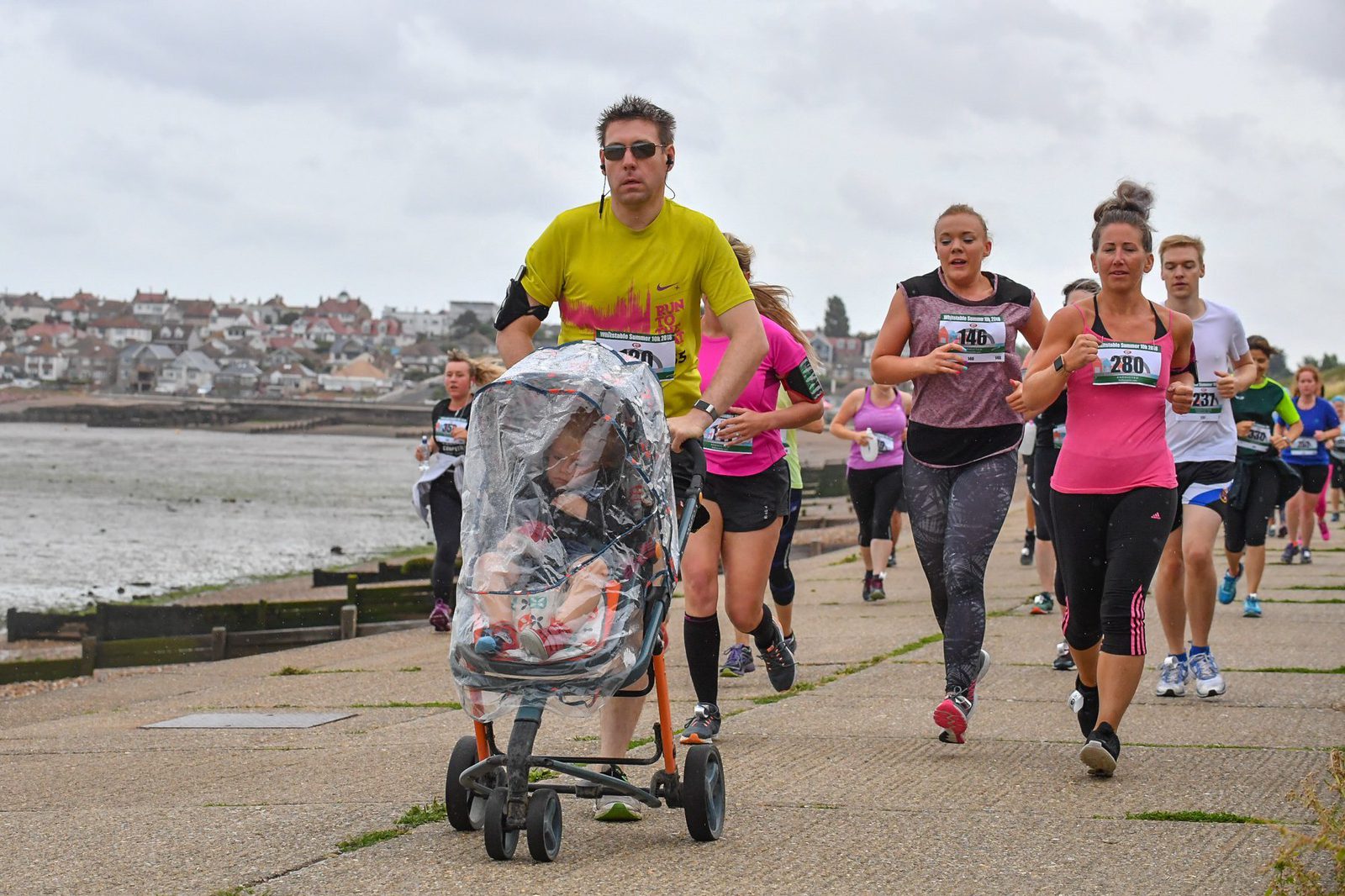 A man in a yellow shirt is pushing a stroller covered in plastic while participating in a race along a coastal path. He is surrounded by other runners wearing race bibs, some in pink shirts. The overcast sky and houses in the background suggest a seaside location.