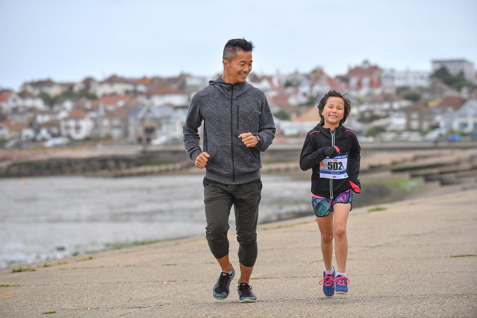 A man and a young girl are jogging together along a seaside path. The man is wearing a dark hoodie and black pants, while the girl is in a black jacket and colorful shorts with a race bib number 502. Buildings and a cloudy sky are visible in the background.