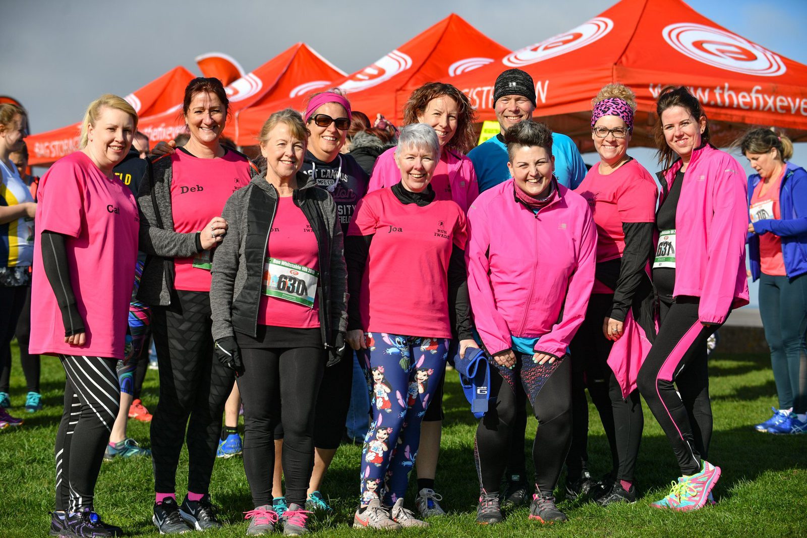 A group of people wearing pink shirts and athletic gear pose together for a group photo on a grassy area during an outdoor event. Red tents with white logos are visible in the background under a clear sky. Various participants are smiling and holding numbered bibs.