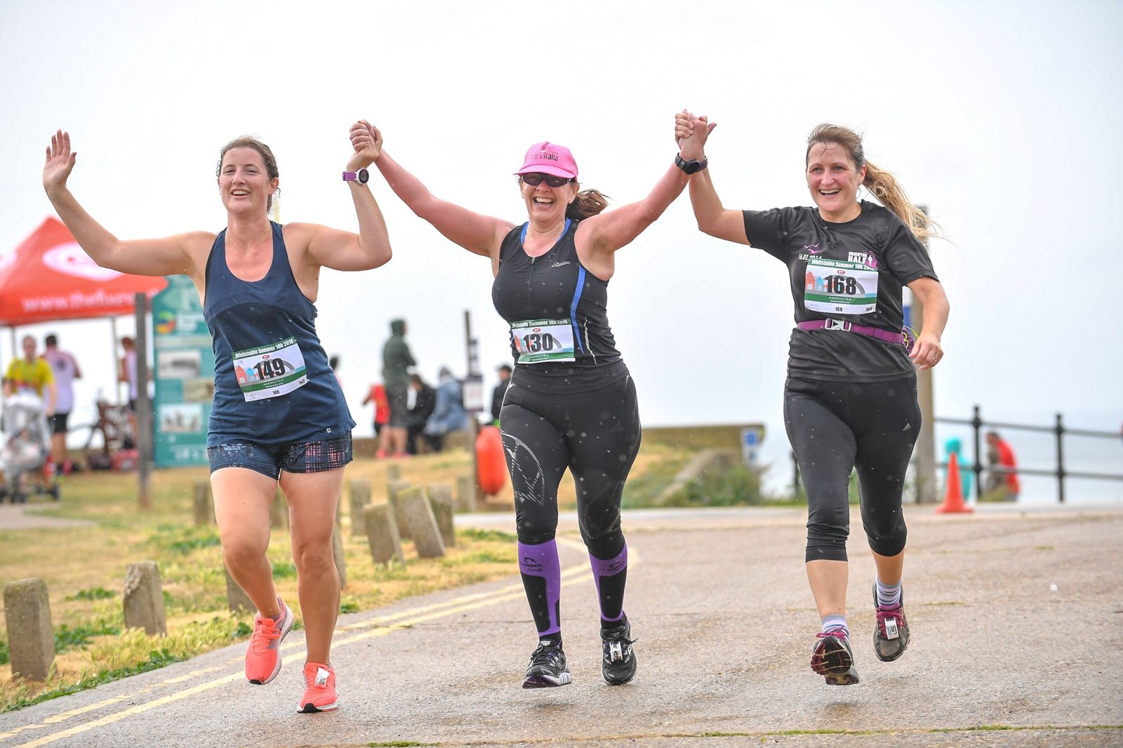 Three women in athletic gear are running outdoors, holding hands and smiling. They each have race numbers: 149, 430, and 168, pinned to their shirts. The setting appears to be a race event by the seaside, with other participants and a red tent visible in the background.