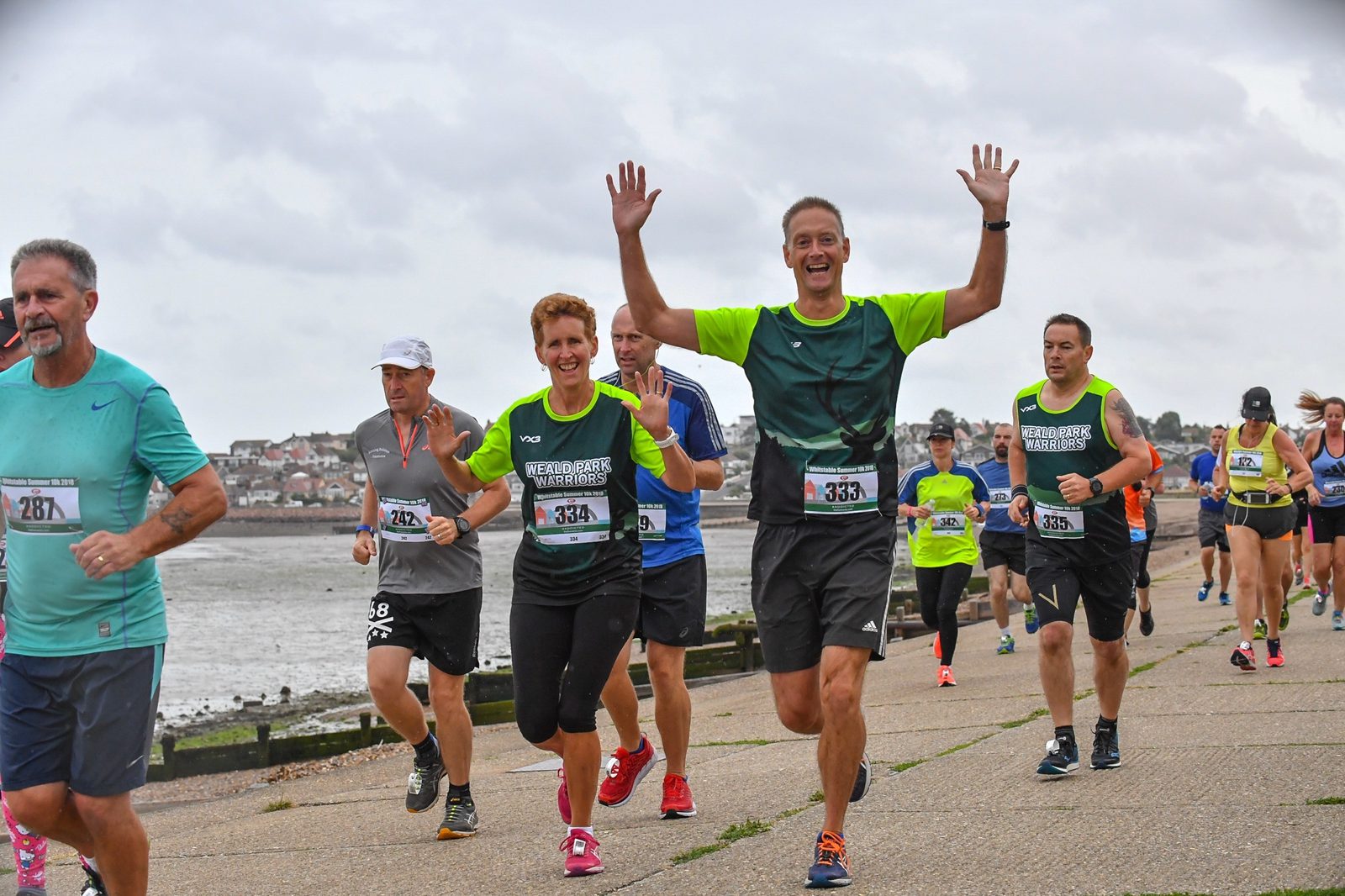 A group of runners participating in a race along a seaside path. One man in a lime green and dark green shirt runs with his arms raised, smiling. Other runners in various athletic wear and race bibs are visible, with the sea and town buildings in the background.