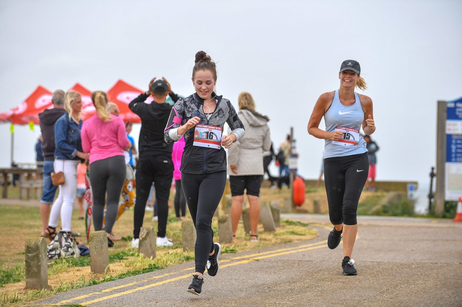 Two women are running on a path during a race, wearing numbered bibs. The woman in front wears black leggings and a jacket, while the one behind her wears black leggings and a gray tank top. A group of spectators and tents are visible in the background.