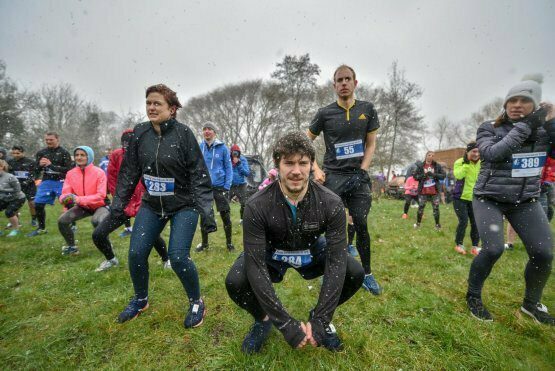 A group of people wearing athletic clothing participate in an outdoor exercise routine in snowy weather. Some are squatting while others stand. Trees and more participants are visible in the background. Many wear race bibs with numbers.