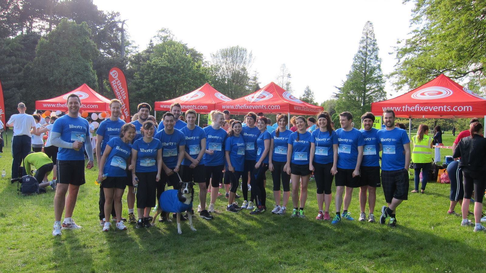 A group of people wearing matching blue shirts pose together on a grassy field. Behind them are red tents with logos. Trees and more people are visible in the background under a bright sky.