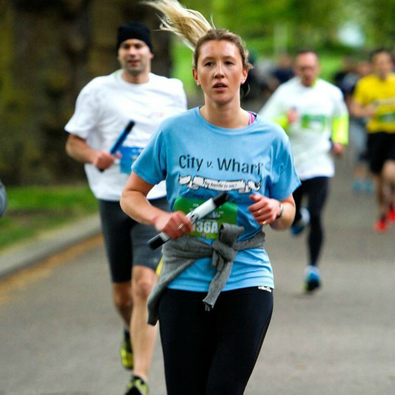 A woman with blonde hair in a ponytail is running in a race, wearing a light blue "City v. Wharf" shirt and black leggings, holding a baton. Other runners are visible in the background, some wearing white shirts. The race appears to be taking place on a road with greenery.