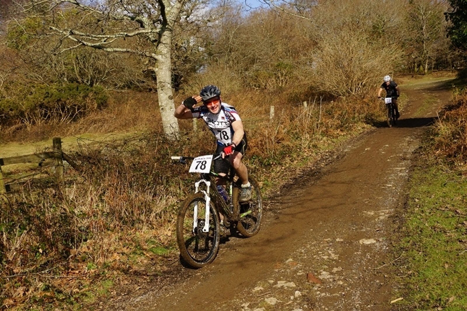 Two cyclists are riding mountain bikes on a dirt trail through a wooded area. The cyclist in the foreground, wearing a helmet and number 78, smiles and raises one hand in a thumbs-up gesture. The second cyclist follows behind on the path.