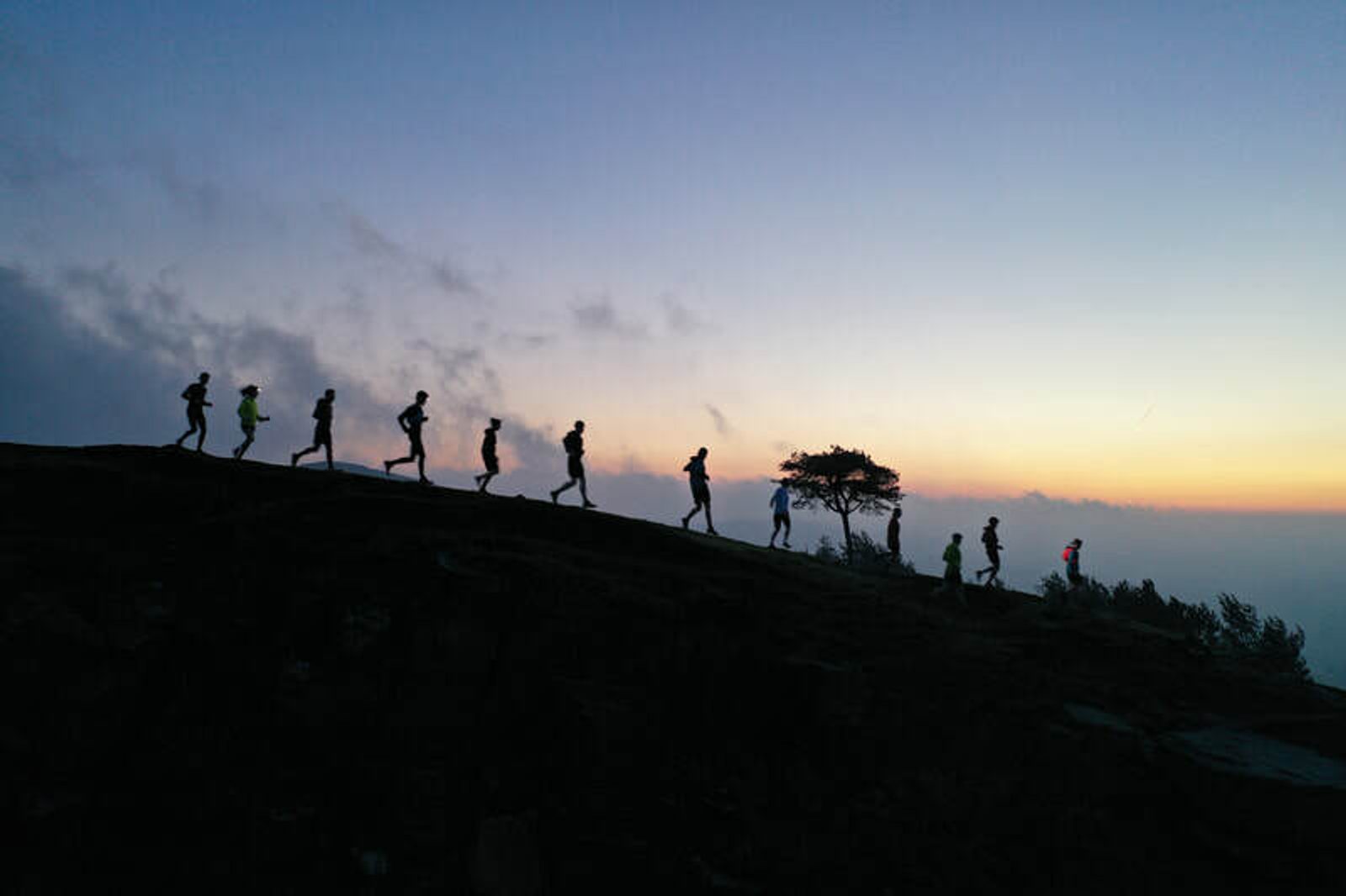 Silhouetted runners in a line jog along a hilltop against a backdrop of a twilight sky. The sky transitions from light to darker blue hues, with soft clouds and a few scattered trees adding depth to the scene.