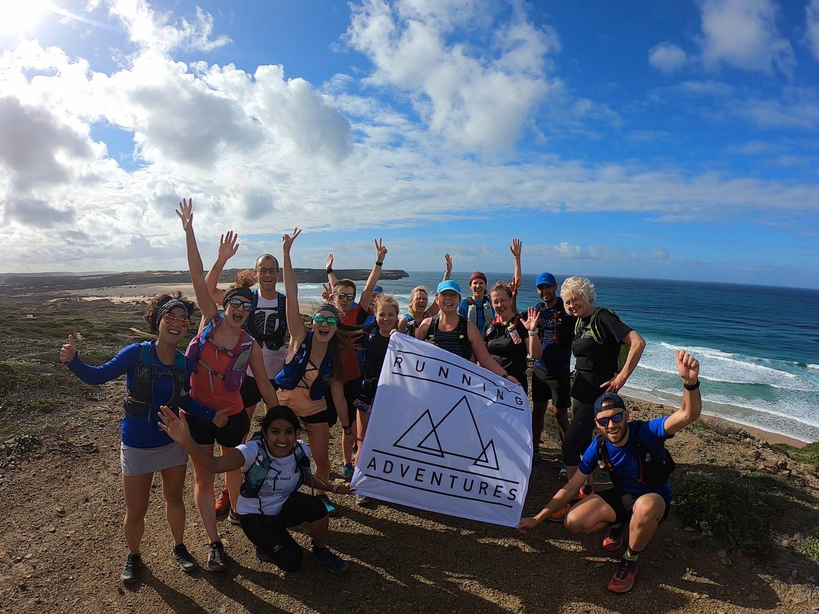 A group of runners poses with a "Running Adventures" banner on a coastal trail. They are smiling and raising their arms in celebration. The background features a scenic ocean view and a partly cloudy sky.