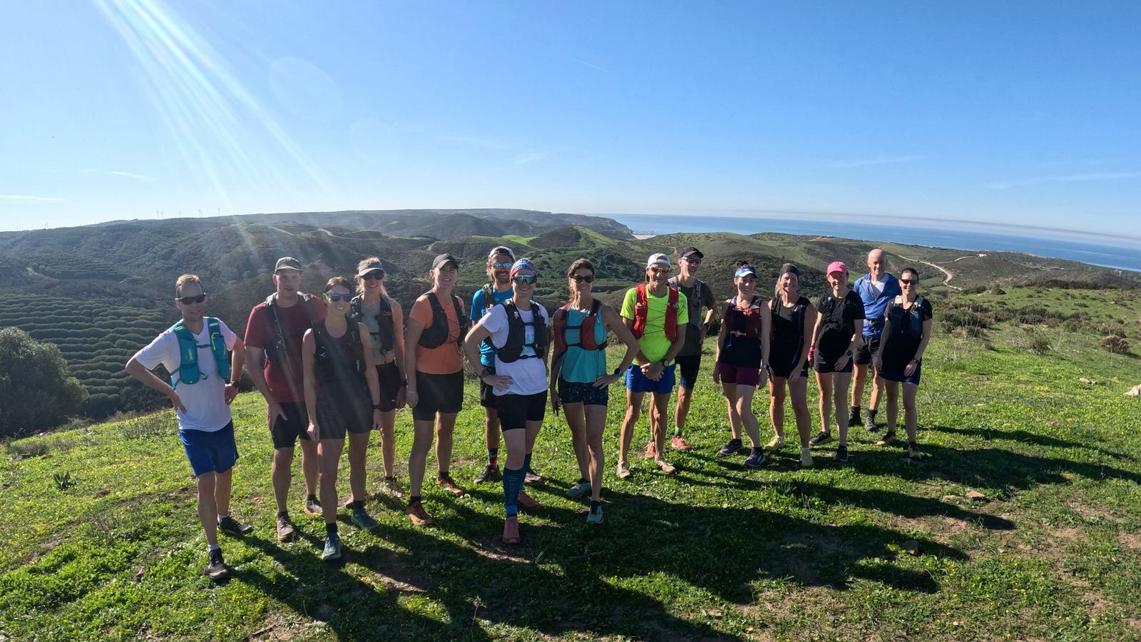 A group of people in athletic wear pose on a grassy hill with a scenic backdrop of rolling hills and the ocean under a clear blue sky. The sun is shining brightly.