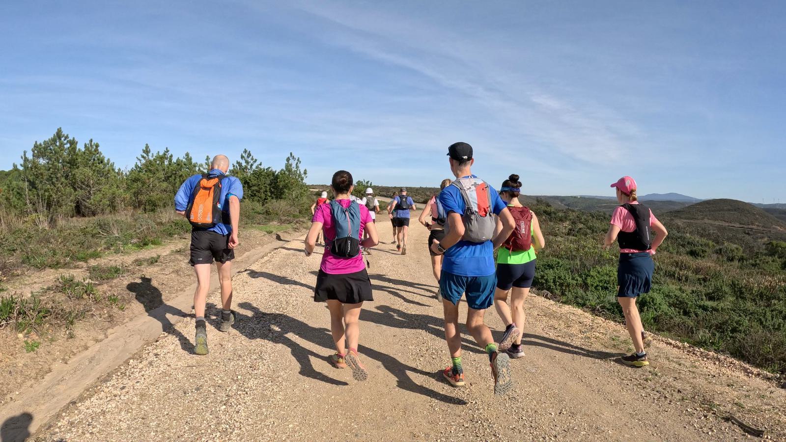 A group of people running on a dirt trail in a rural area, surrounded by greenery and small hills. They are wearing athletic gear and backpacks. The sky is clear and blue, indicating a sunny day.