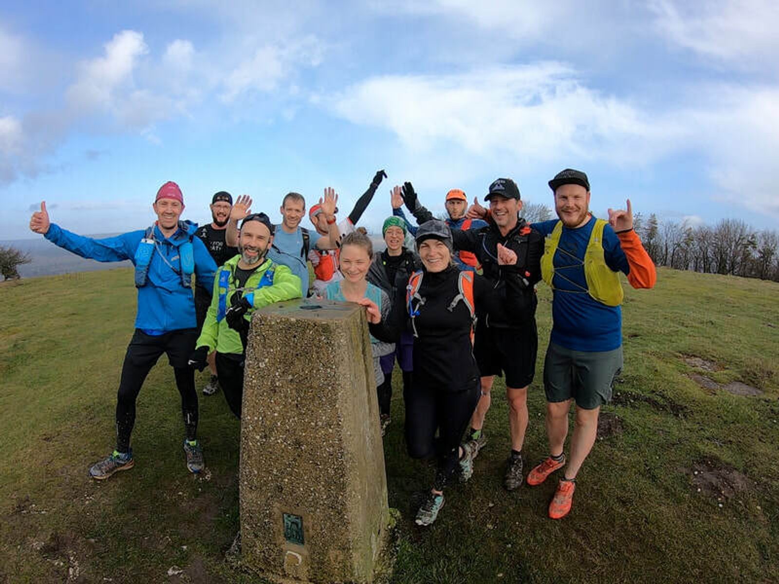 A group of eleven smiling people, dressed in outdoor athletic gear, pose around a concrete marker on a grassy hilltop. Some give thumbs up, and the sky is partly cloudy. The group appears enthusiastic and happy, suggesting a successful hike or run.