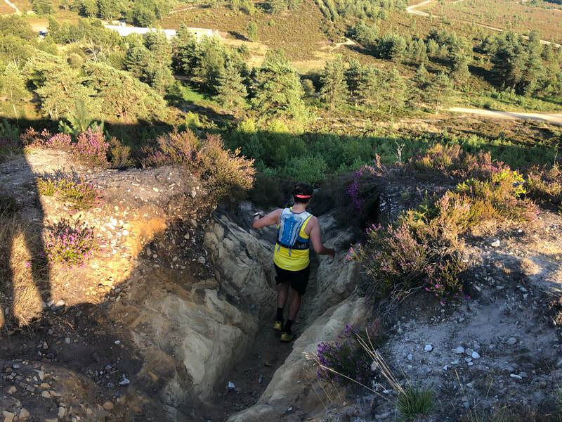A person wearing a yellow top and a hydration pack is navigating a rugged, eroded path in a hilly, forested area. The terrain is challenging with steep, narrow sections and patches of vegetation. Pine trees and a winding road are visible in the background.