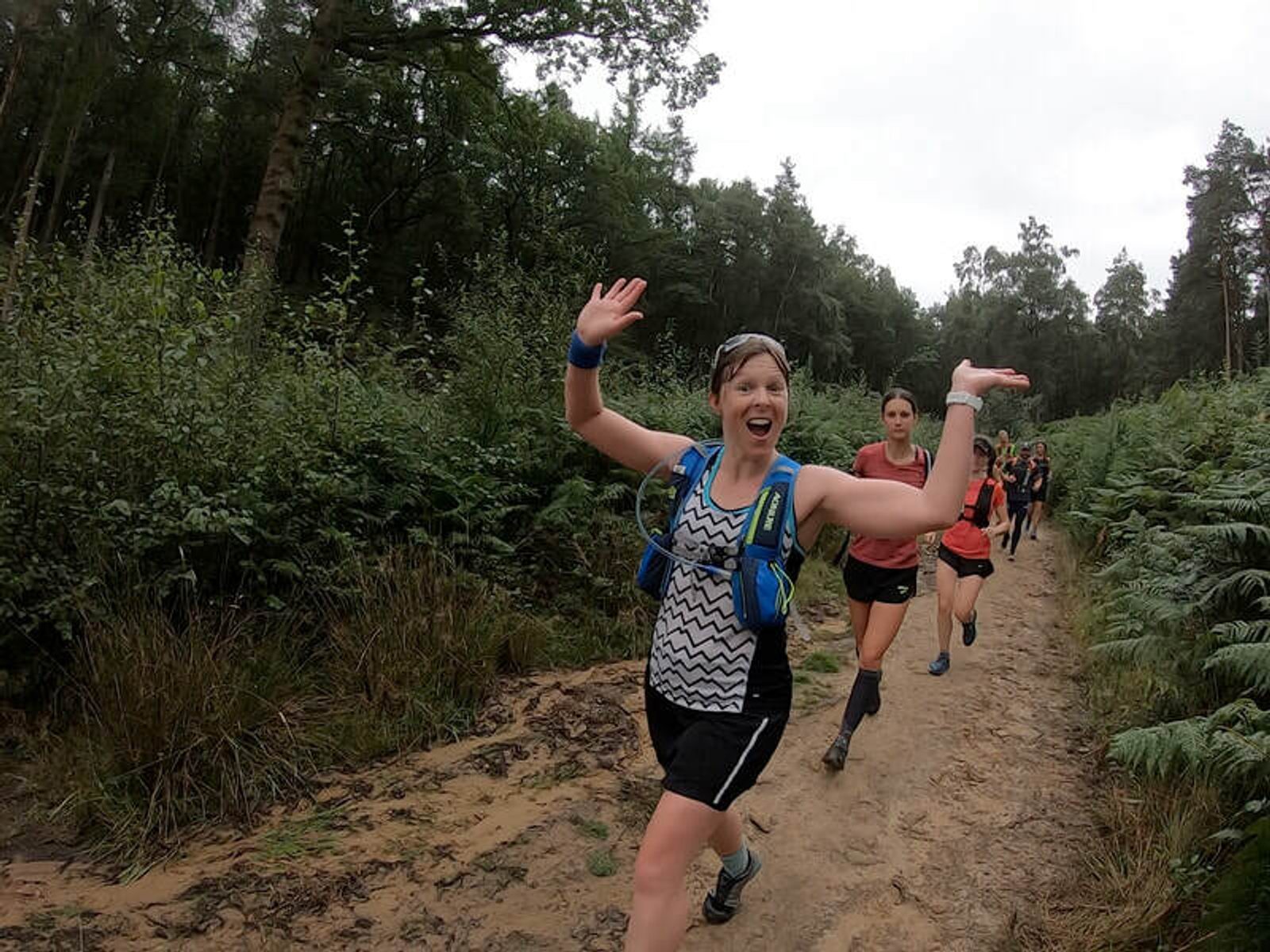 A woman wearing a blue hydration pack and patterned tank top runs on a dirt trail through a forest, smiling and waving at the camera. Other runners follow behind her, surrounded by greenery and trees.