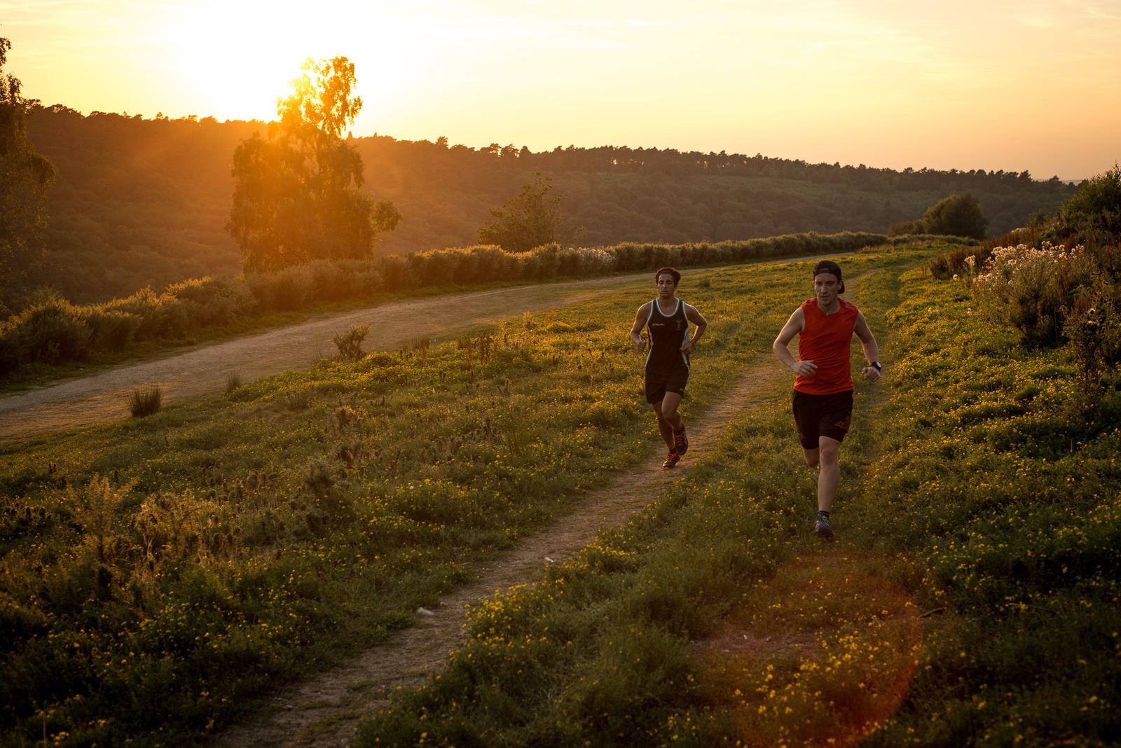 Two people jogging on a dirt path through a grassy field at sunset. The sun is low in the sky, casting a warm, golden light on the landscape. Trees and rolling hills are visible in the background. The joggers wear athletic clothing, one with a dark top and the other in a red top.