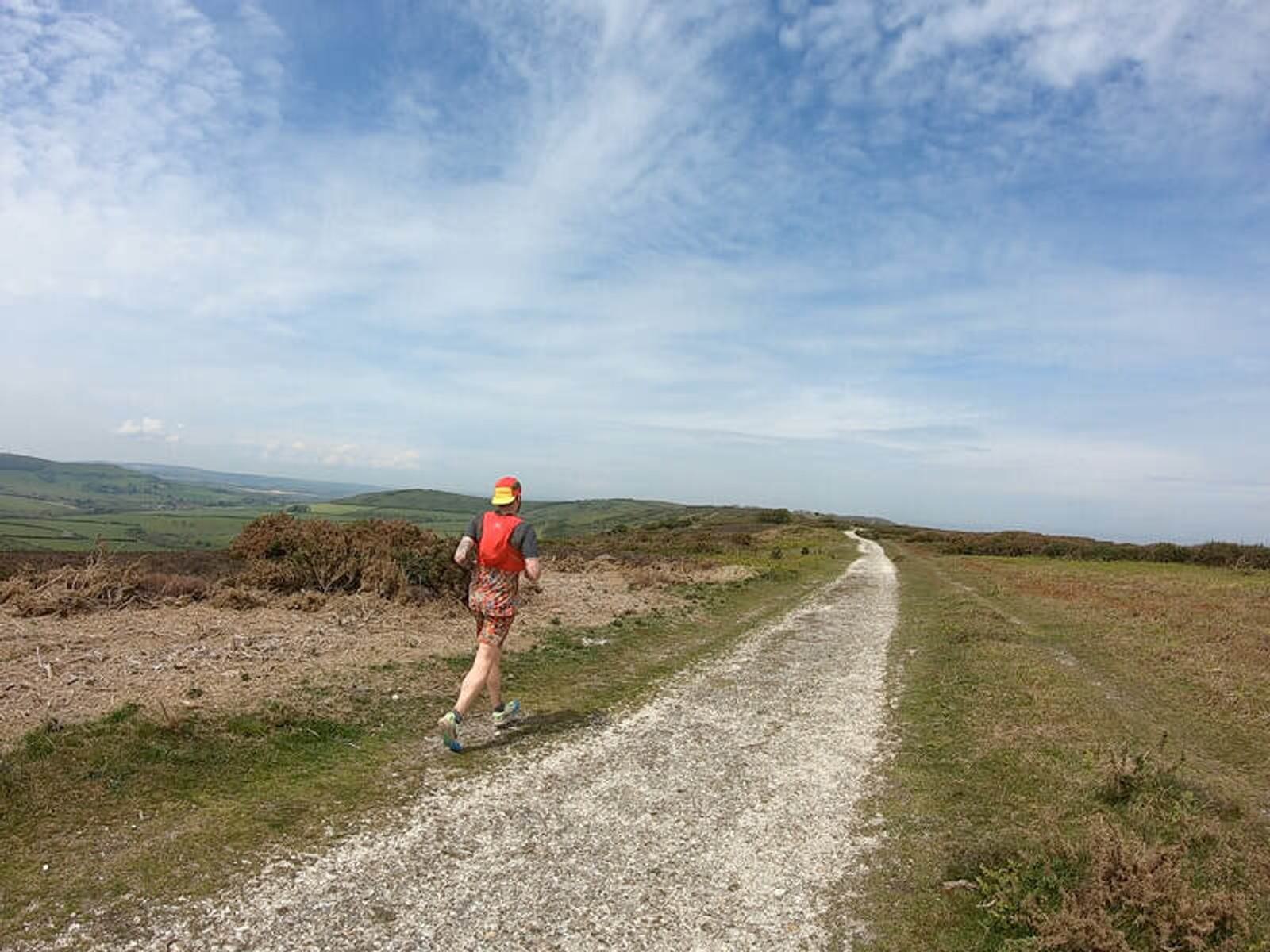 A person in a red tank top, shorts, and a bright cap is running along a gravel path through a grassy, hilly landscape under a blue sky with scattered clouds. The path extends far into the distance, surrounded by fields and low-growing vegetation.