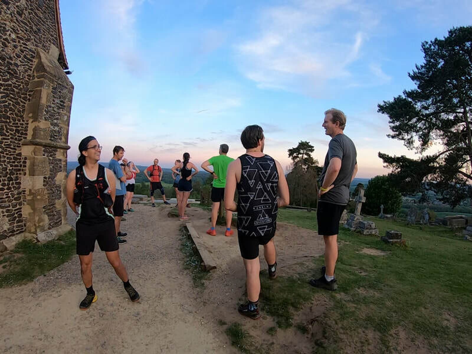 A group of people wearing athletic clothing gather outside a stone building during a picturesque sunset. Some are standing and chatting, while others look into the distance. The background includes a tree, rocky terrain, and an expansive sky with light clouds.