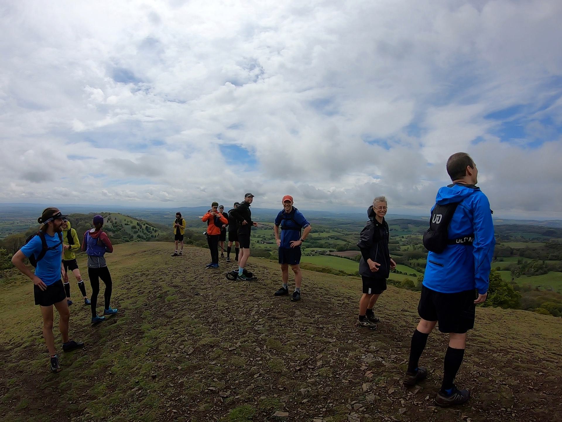 A group of people wearing athletic gear stand on a grassy hilltop with a scenic view. The sky is overcast, and the landscape below includes fields and forests. Two people in the foreground are using Nordic walking poles.