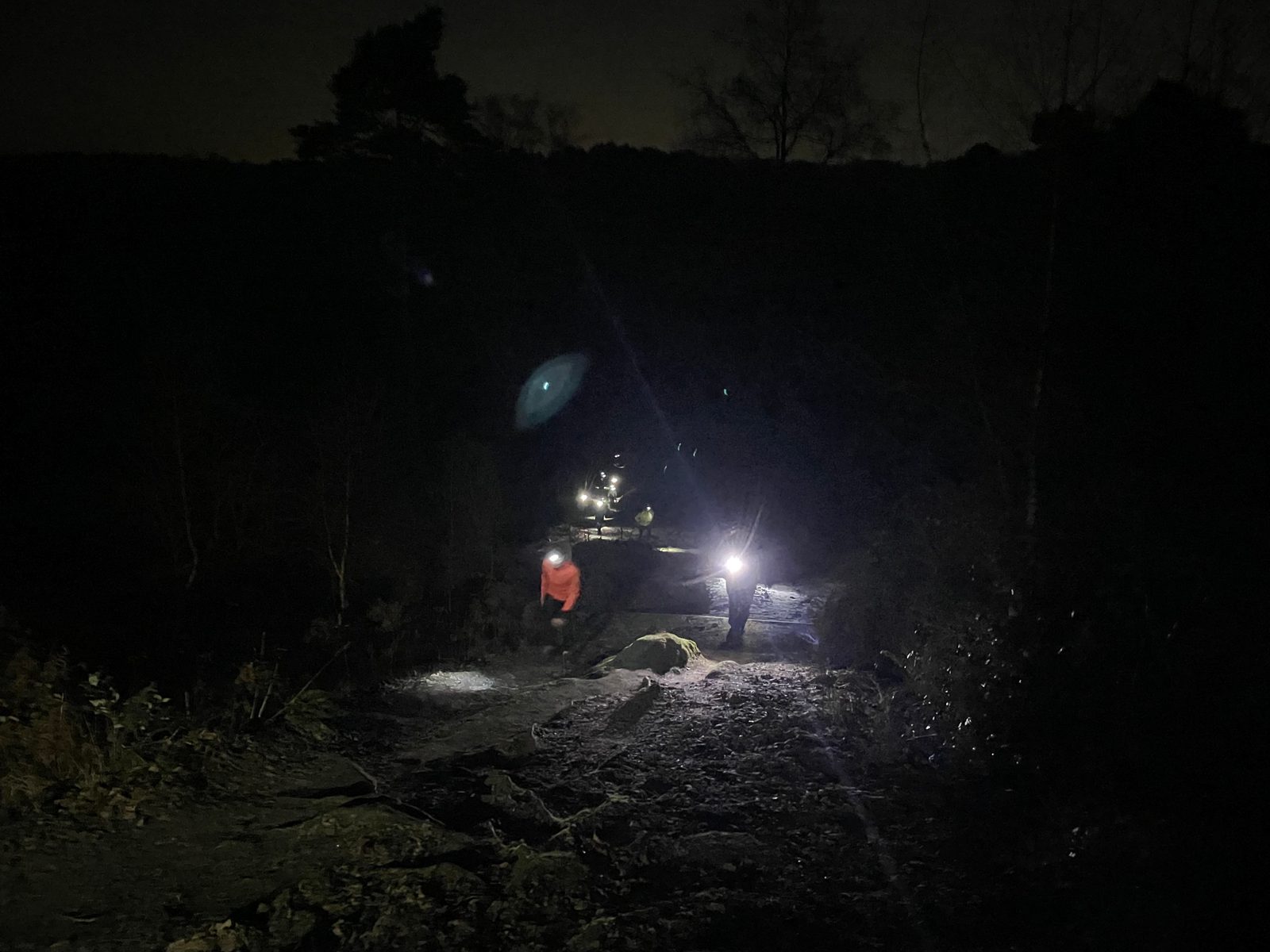 Several people wearing headlamps and reflective jackets walk along a rocky trail at night, with their lights illuminating the dark path and surrounding trees.