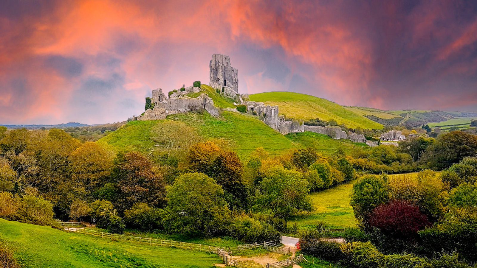 A picturesque scene of a hillside castle ruin surrounded by lush green rolling hills and dense foliage. The sky above is dramatic with vibrant orange and pink hues, suggesting either a sunset or sunrise, creating a striking and colorful backdrop for the medieval structure.