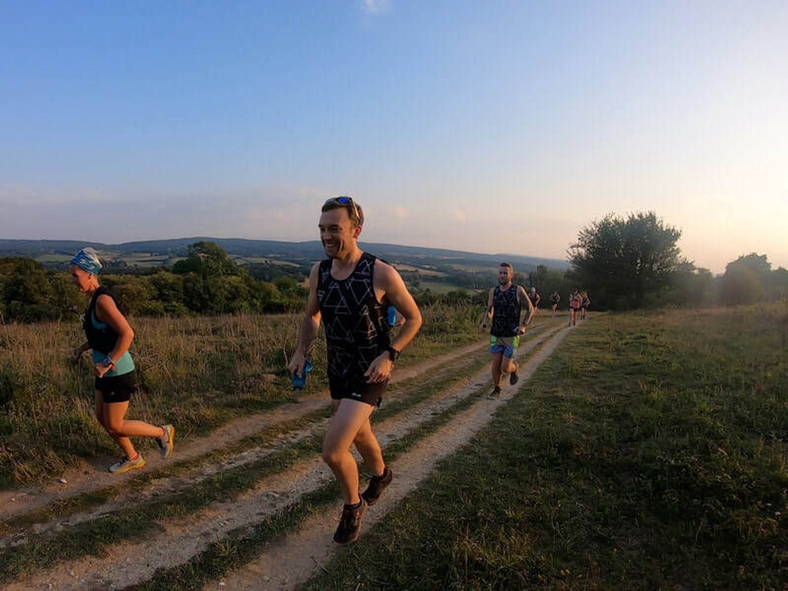 People are running on a grassy trail during sunset. The landscape is open with hills and trees in the background. The runners wear casual athletic gear and appear to be enjoying the outdoor activity.