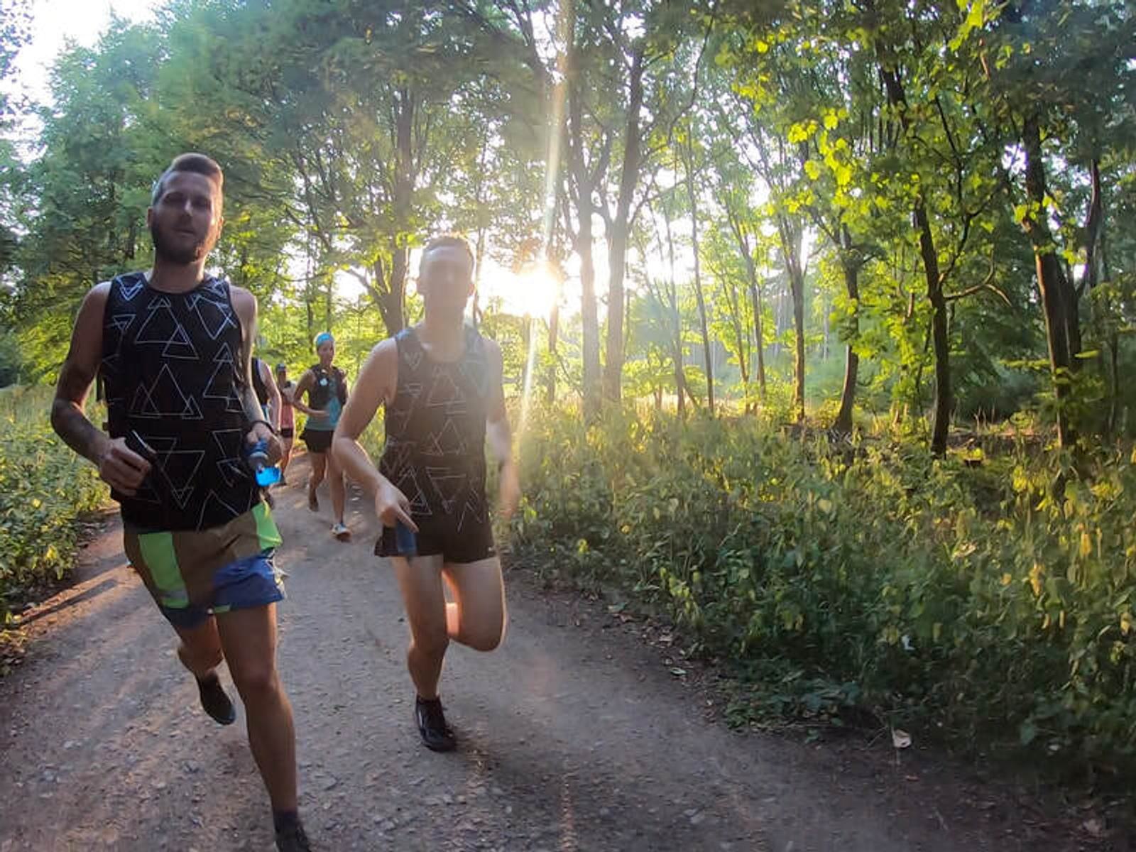 Four people are jogging on a dirt path through a forest during sunset. The sun is shining through the trees, creating a warm, dappled light. They are wearing athletic gear and appear focused and energetic.