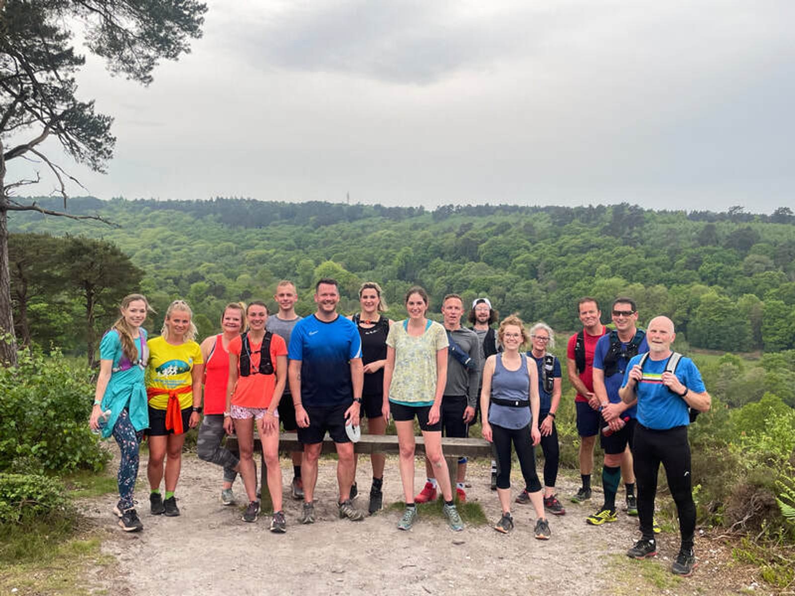 A group of people wearing casual athletic clothing are standing on a trail in front of a lush green forest. The sky is cloudy. They are smiling and appear to be enjoying an outdoor activity.