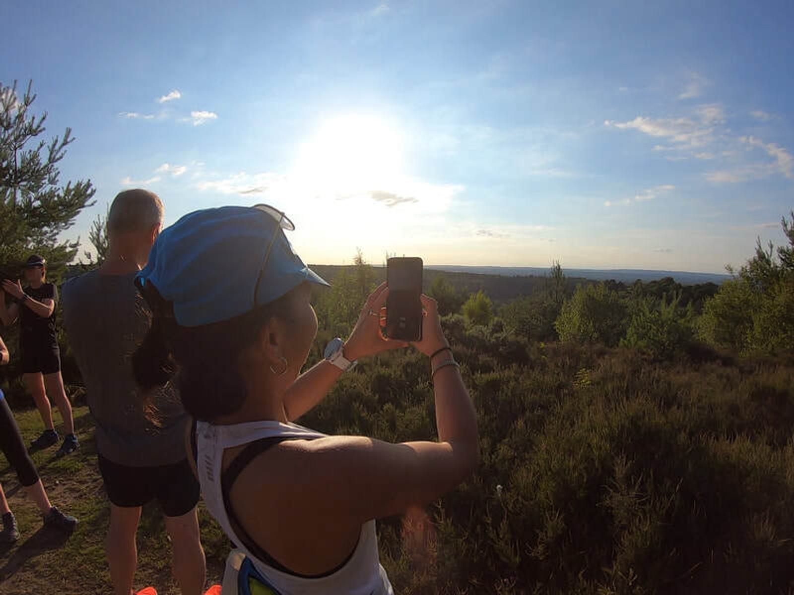 A woman wearing a cap takes a photo with her phone. She stands on a grassy hill surrounded by trees at sunset, with a man and others nearby. The sky is blue with a bright sun, casting a warm glow over the landscape.