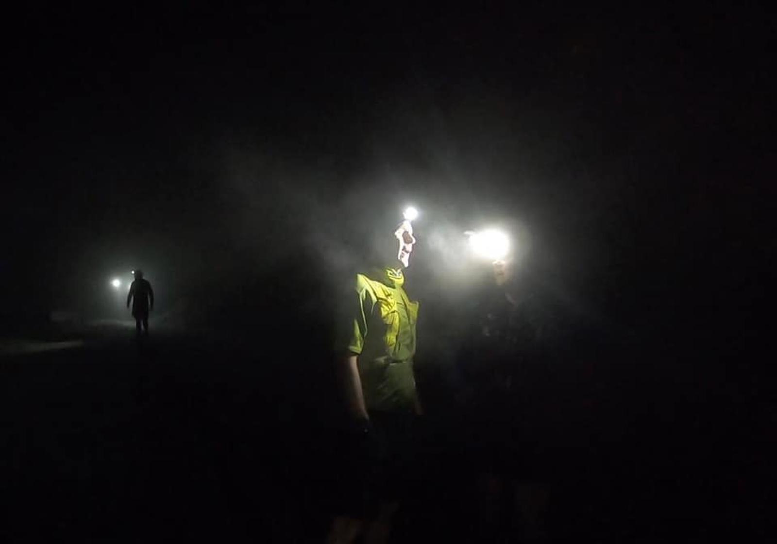 Two people wearing headlamps navigate through a dark, foggy environment. A faint silhouette of another person is visible in the background, adding to the mysterious atmosphere.