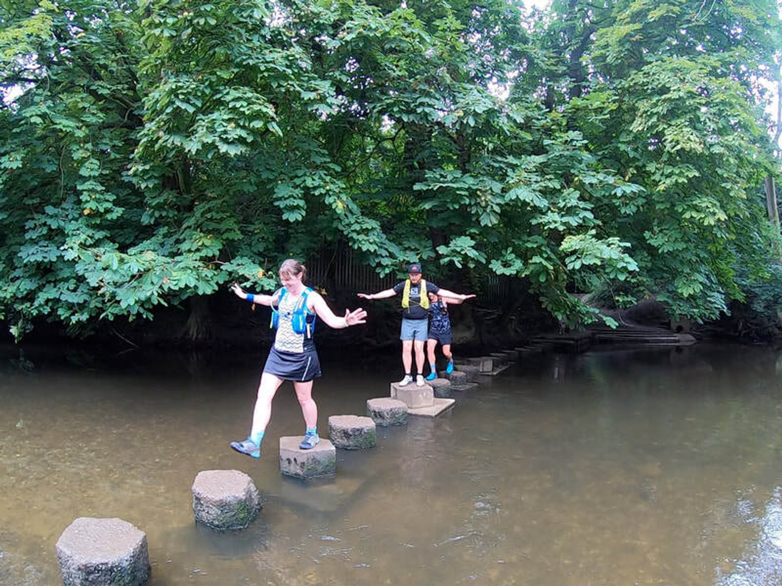 People crossing a shallow stream on stepping stones, surrounded by lush green trees. They appear focused and balanced as they make their way across.