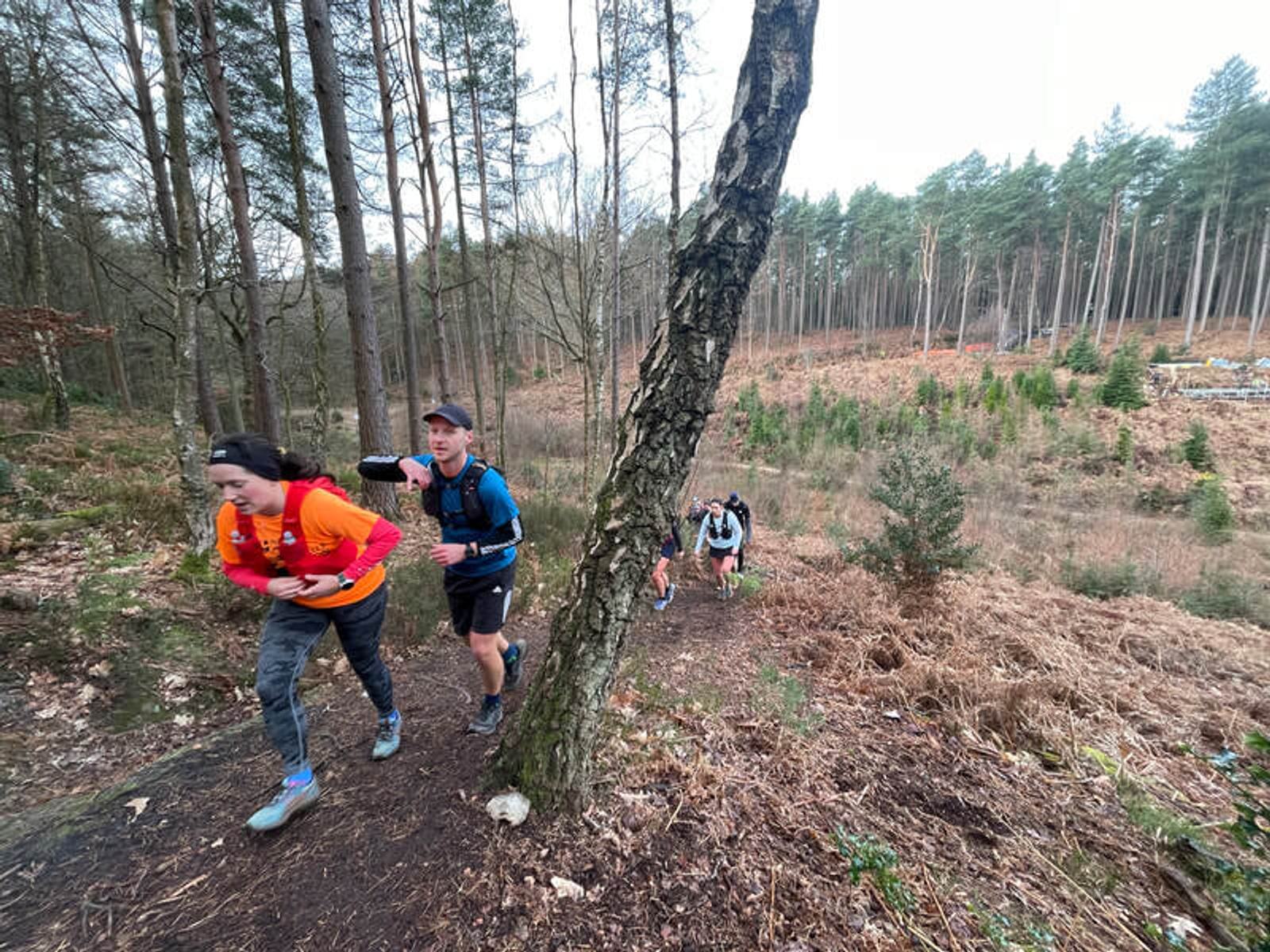Two people in athletic gear run up a hill in a forested area, with two more runners in the background. The scene is surrounded by trees and dry foliage on a clear day.