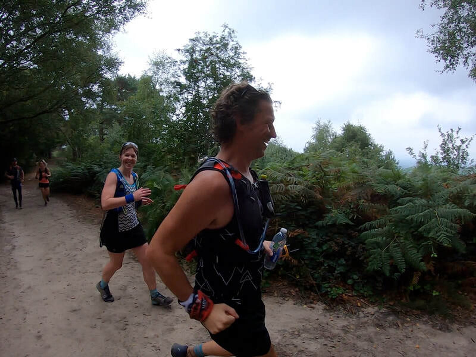 Two people are running on a forest trail, surrounded by greenery. They are wearing athletic gear, and the day appears overcast. Both are smiling and enjoying the run.
