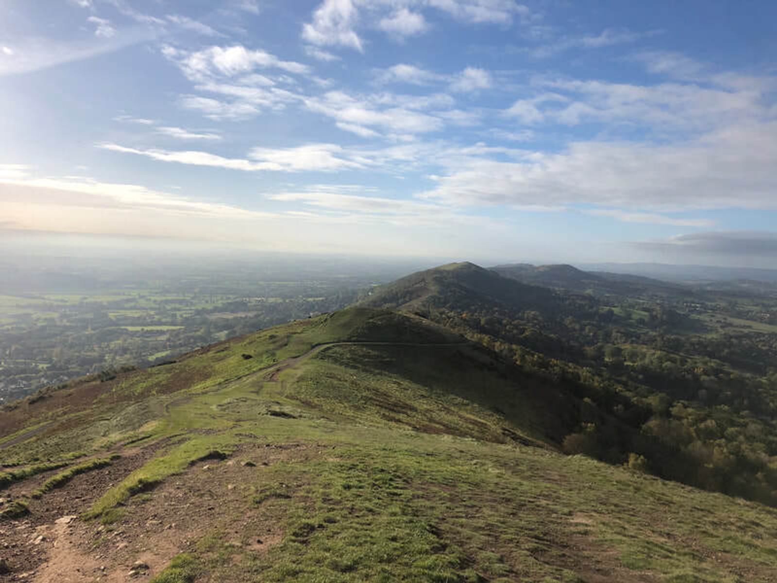 A scenic view of a green ridge stretching into the distance under a blue sky with scattered clouds. The landscape below is a mix of fields and forested areas, with a distant horizon.