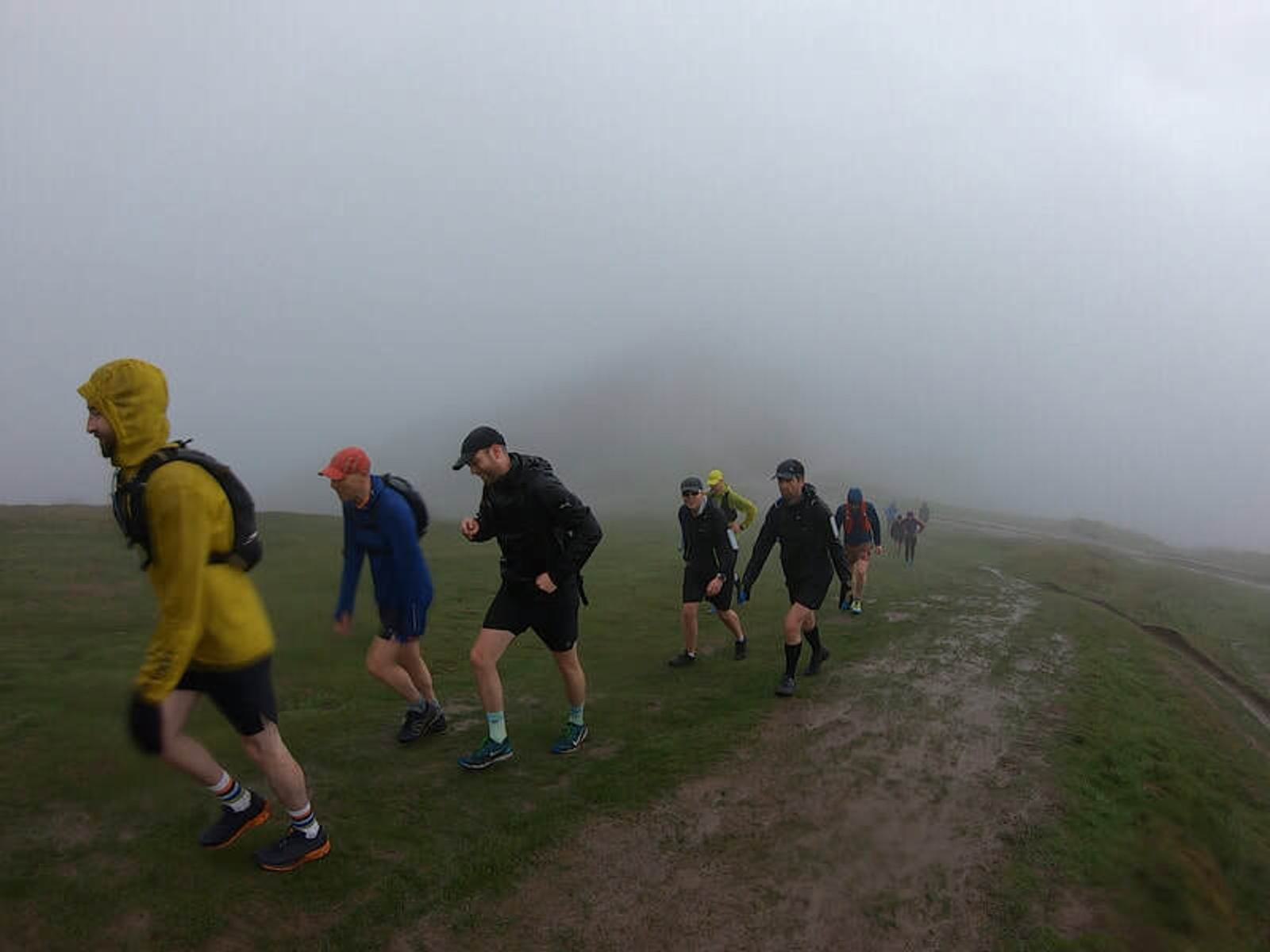 A group of people, dressed in rain gear, hike up a misty, grassy hill. The sky is overcast, and the ground appears wet and muddy, creating a foggy atmosphere.