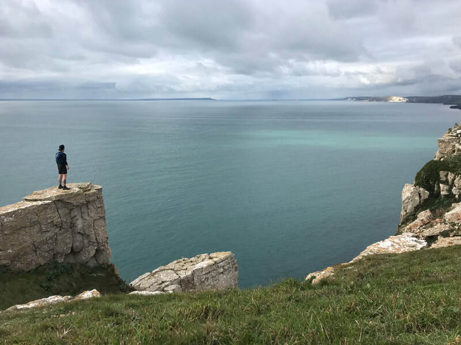 A person stands on a rocky cliff edge, overlooking a vast body of water beneath a cloudy sky. The scene is tranquil, with expansive sea views and distant white cliffs.