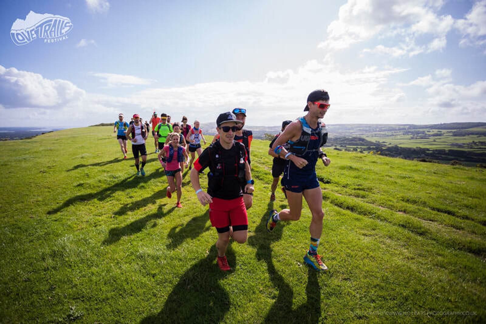 A group of runners in colorful athletic gear participates in a trail race on a grassy hillside under a partly cloudy sky. The backdrop features a scenic view of fields and distant hills.