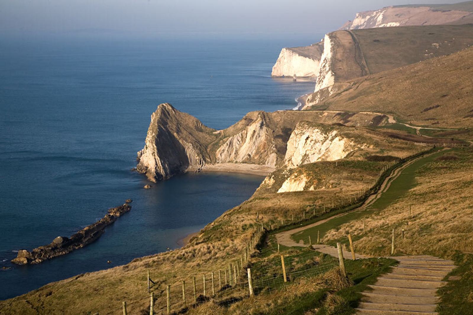 A scenic coastal view featuring dramatic cliffs and a natural arch jutting into the sea, surrounded by calm blue waters and grassy hills. A walking path winds along the hilltop, offering panoramic views of the rugged coastline.