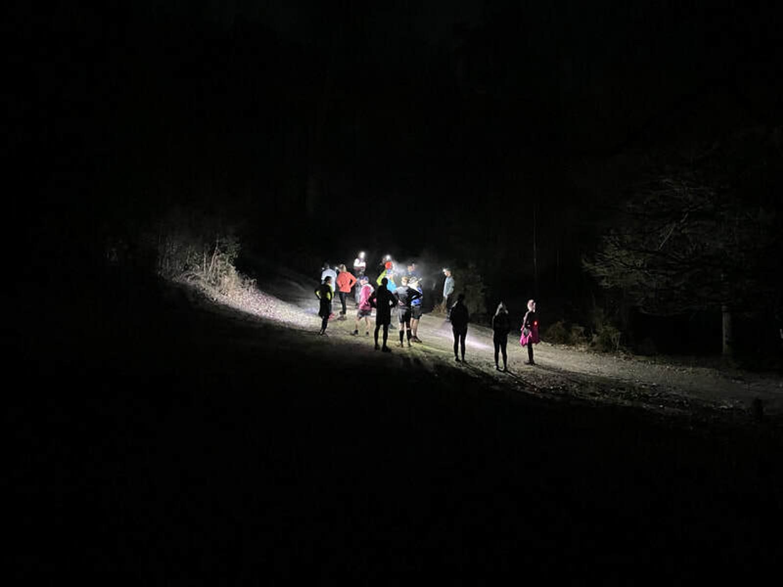 A group of people gathered at night on a dimly lit path, surrounded by darkness. They are illuminated by a bright light source, creating silhouettes against the backdrop of trees. The scene conveys a sense of adventure or exploration.