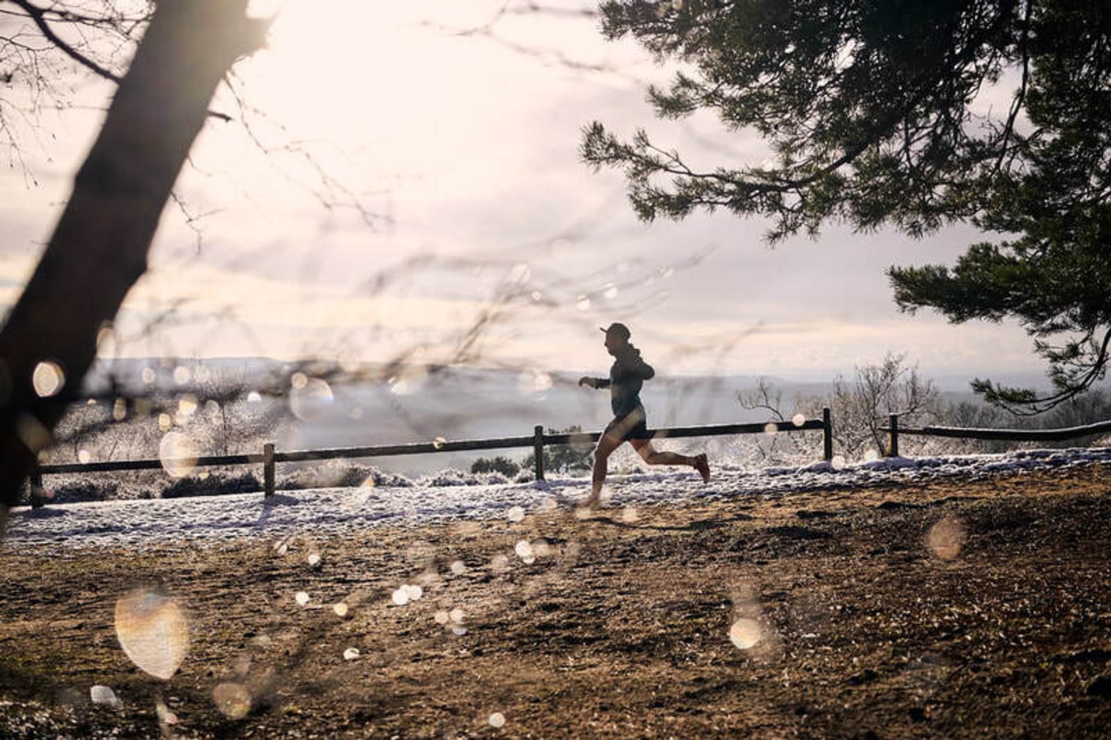 A person jogs along a snowy path under a cloudy sky, surrounded by bare trees. Glowing light reflections and blurred branches are visible in the foreground, creating a serene winter atmosphere.