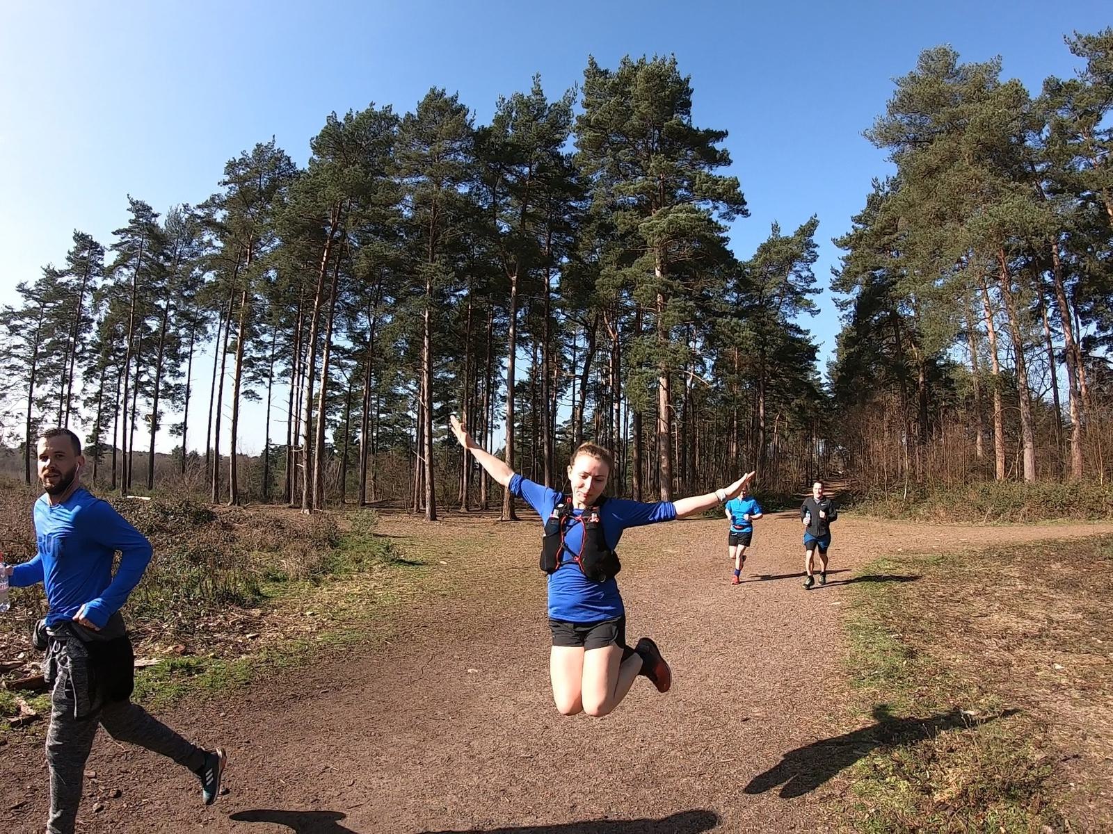 A woman in a blue shirt jumps joyfully on a forest trail, surrounded by tall trees under a clear blue sky. Three other runners in blue can be seen on the path behind her.