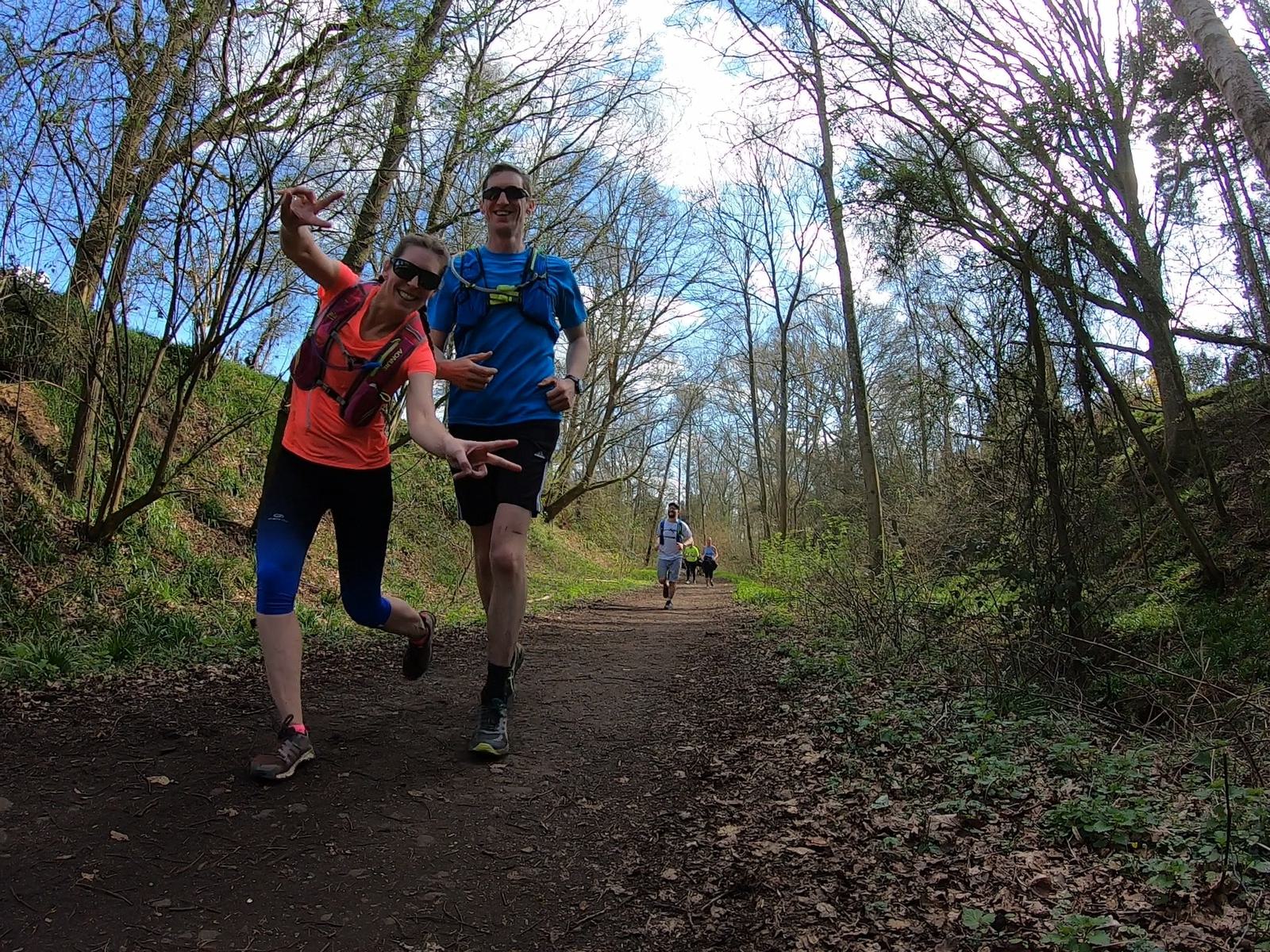 Two people jogging on a forest trail, smiling and gesturing playfully at the camera. They are wearing sunglasses and athletic gear. The path is surrounded by trees and greenery, with a couple of runners in the background.