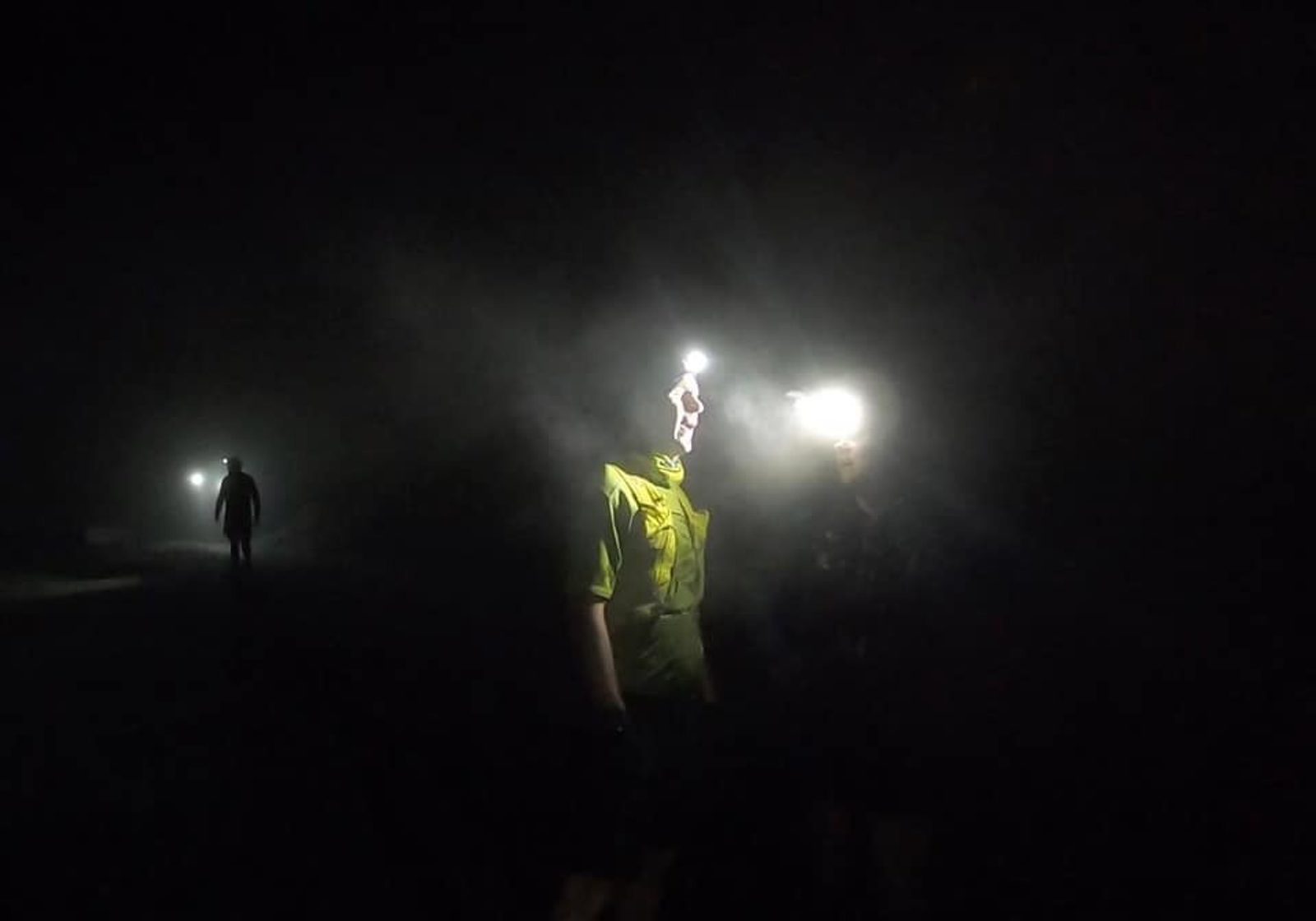 Three people are outdoors in the dark, wearing headlamps that shine brightly through the darkness and mist, illuminating their faces and creating a glowing effect in the night.
