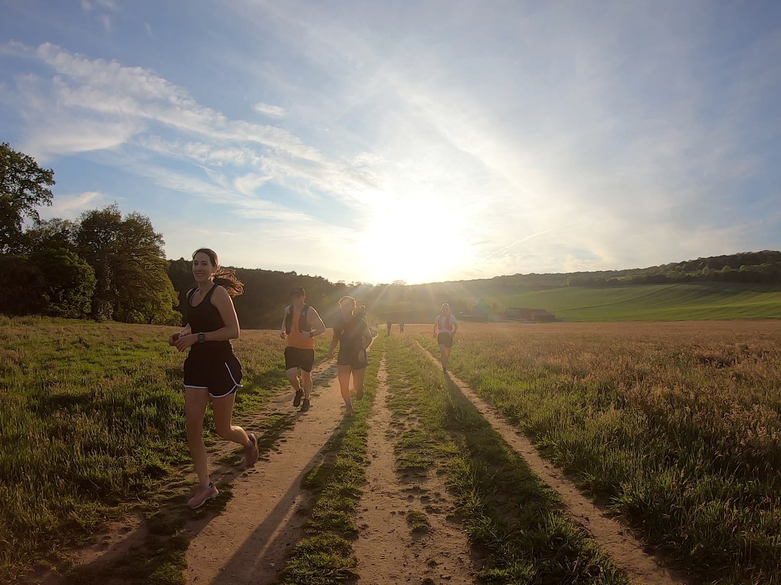 A group of people jogs on a dirt path through a grassy field during sunset. The sun is low in the sky, casting long shadows. Trees and hills are visible in the background under a partly cloudy sky. The lead runner is wearing a black tank top and shorts.