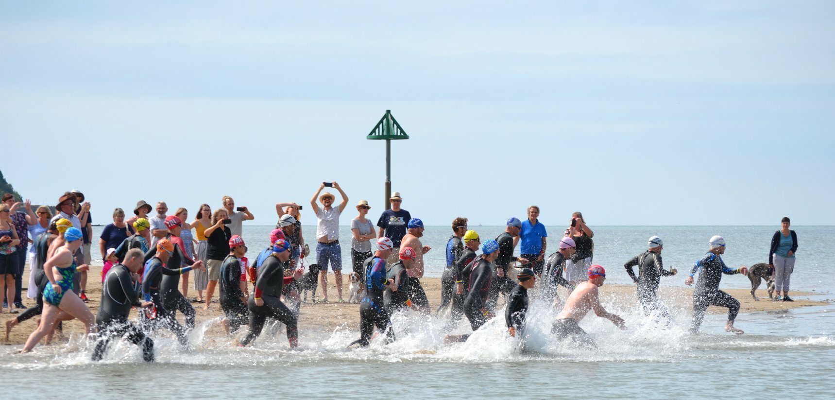 A group of people in wetsuits and swim caps rush into the ocean to start a race. Spectators stand on the beach, some taking photos, cheering, and watching. The sky is clear, and there is a triangular marine beacon in the background.