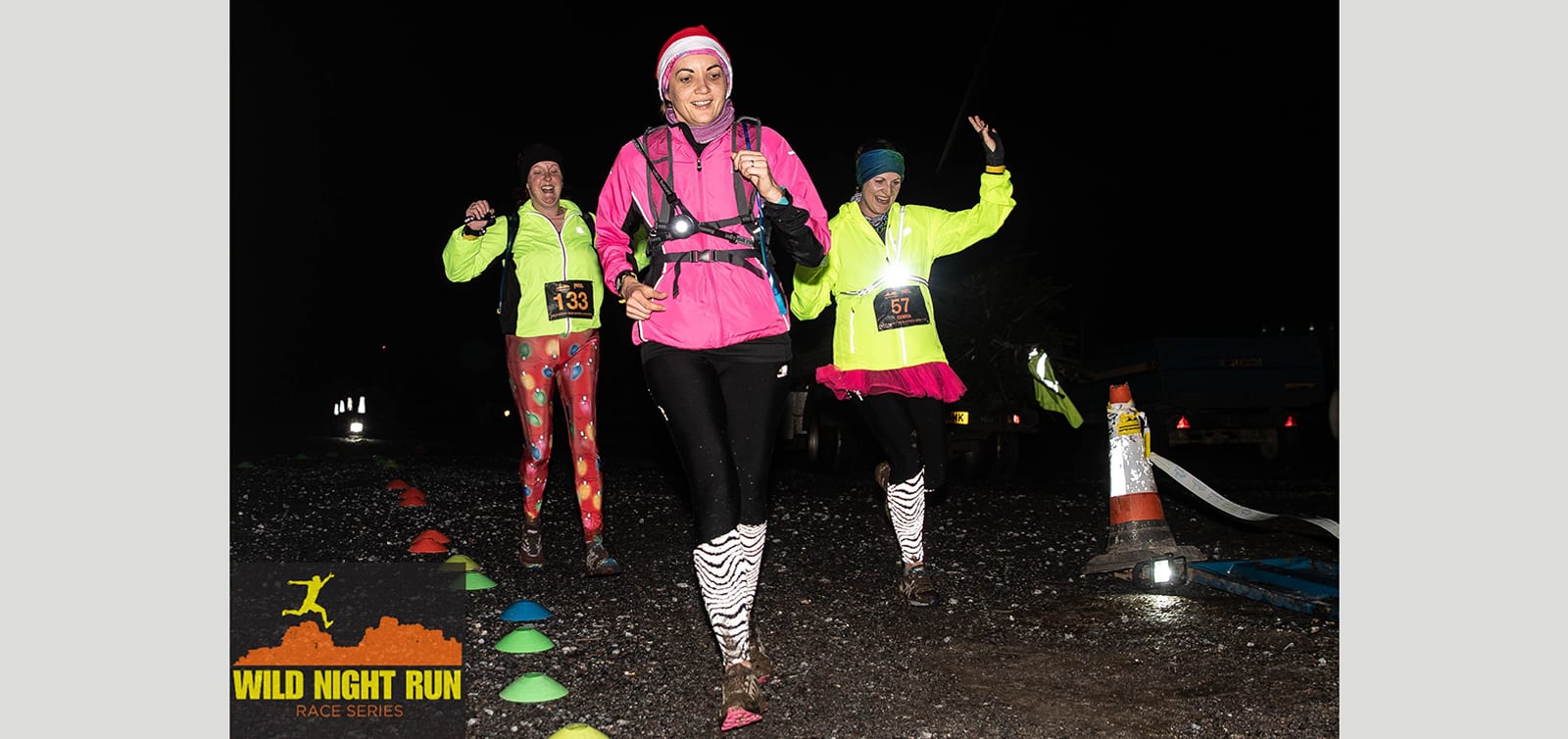 Three runners participate in a night race wearing headlamps, bright clothing, and race numbers 133, 157, and 163. The runner in the center wears a Santa hat and pink jacket. The Wild Night Run logo is visible in the bottom left corner.