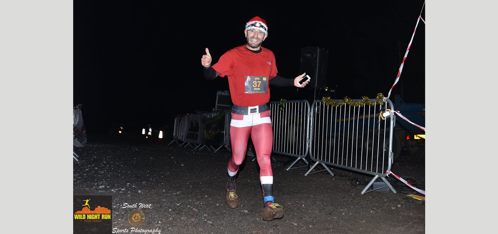 A runner in red pants and a red shirt, wearing a headlamp, smiles and gives a thumbs up as he races at night. Barriers and caution tape line the path behind him. He wears a race bib with the number 37. The background is dark, and there are some decorations visible.
